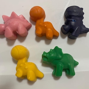 Könnte beinhalten: Fünf bunte, dinosaurierförmige Wachsmalstifte in Rosa, Orange, Marineblau, Gelb und Grün. Die Wachsmalstifte sind auf einer weißen Oberfläche angeordnet. Die Dinosaurier umfassen einen Stegosaurus, einen Brontosaurus, einen Triceratops und zwei weitere nicht identifizierte Arten.