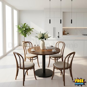 May include: A round dark wood dining table with a black pedestal base, surrounded by four wooden chairs with light beige upholstered seats. A white vase with white flowers, two tall thin candles, and a stack of books sit on the table surface.