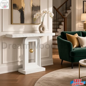 May include: A white console table with a faux marble top and base, featuring a central arch-shaped support structure with a suspended gold sphere. The table has a rectangular surface and a solid, tiered base, providing a clean, structural design.