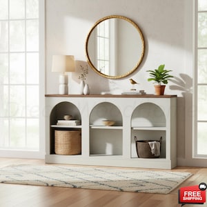 May include: A white console table with three arched open shelves, a brown wooden top, and a round gold-framed mirror. The shelves contain a woven basket, a dark brown basket, books, and bowls. A white lamp and a green plant sit on the top surface.