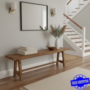 May include: A wooden bench with a rectangular top and angled legs. The bench is in a hallway with a mirror, wall sconces, and a staircase. Beige towels are stacked on the bench, and a vase of dried flowers sits nearby.