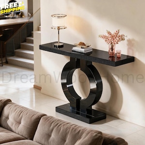 May include: A black console table with a rectangular top and a base featuring two large circular supports. The table surface holds a cylindrical lamp with a white shade, a stack of books, a small decorative bowl, and a glass vase with flowers.