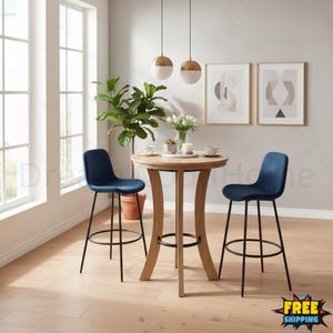 May include: A round wooden pub table with a light oak finish and three curved legs, paired with two navy blue velvet bar stools featuring black metal frames and circular footrests. The table is set with white ceramic cups and a vase of flowers.