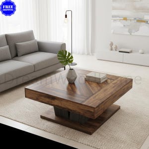 May include: A square coffee table with a dark brown wood grain finish featuring a chevron pattern on the tabletop. The table has a thick frame, a solid base, and a central support structure. Two stacked books and a small vase with a green plant sit on top.