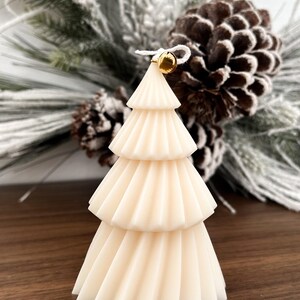 May include: A cream-colored, tiered Christmas tree candle with a gold bell at the top. The candle has a textured, pleated design. The background includes pinecones and frosted greenery, suggesting a holiday theme.