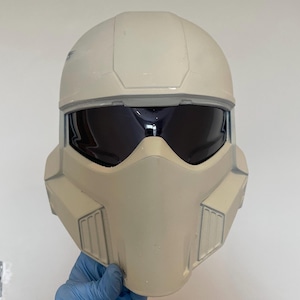 May include: A light beige, futuristic helmet with a dark visor. The helmet has a rounded top and angular features around the face and sides. The visor is dark, and the helmet appears to be made of a hard plastic material. The helmet is held by a hand wearing a blue glove.
