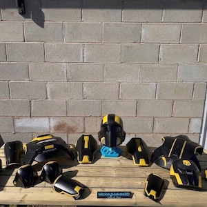 May include: A set of black and yellow 3D-printed armour components, including a helmet, chest plate, and limb guards. The items are displayed on a wooden surface against a brick wall. The text "MickeyMakes3D" is visible.