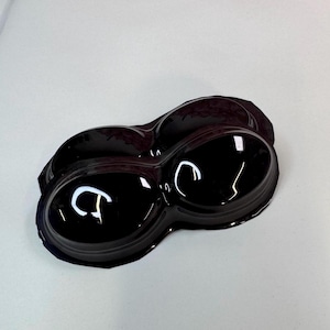 May include: A glossy, dark burgundy plastic container with two connected oval-shaped compartments. The surface reflects light, creating bright white highlights. The container sits on a plain white surface.