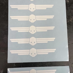 May include: White vinyl decals featuring a skull with wings design. The decals are arranged on a light blue backing sheet. The design is repeated multiple times, with one decal cut separately. Suitable for various applications.