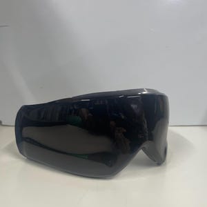 May include: A pair of sleek, wraparound sports sunglasses featuring a single continuous dark black lens that provides full coverage. The frame is constructed from a matte black material, designed with a curved, aerodynamic shape for a secure fit.