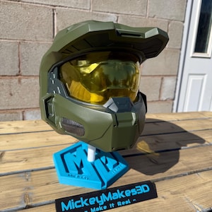 May include: A green and gold helmet, similar to a science fiction character's, is displayed on a blue stand. The helmet has a gold visor. The stand has the text "MickeyMakes3D - Make It Real -" on it. The background is a brick wall.