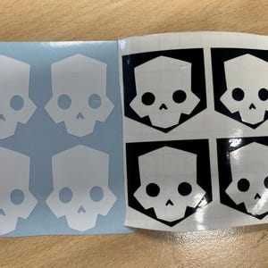 May include: Two sheets of stickers featuring a stylized skull design. One sheet has white skulls on a light blue background. The other sheet has black skulls with white details on a white background. The skull design is geometric.