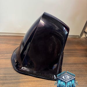 May include: A glossy, dark purple, curved, aerodynamic shell-like object with a smooth, reflective surface. In the bottom right corner, there is a blue, metallic-looking logo featuring the text MICKEY MAKES 3D with a central gear icon.