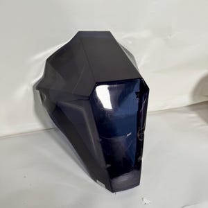 May include: A dark blue, geometric object with a glossy finish. The object has multiple facets, creating a faceted appearance. The shape is reminiscent of a stylized mask or decorative piece.