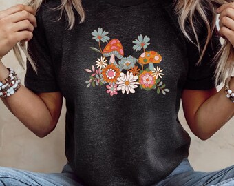 May include: Dark gray t-shirt featuring a colorful floral and mushroom graphic. The design includes orange, pink, blue, and white flowers, along with two mushrooms with orange and white polka dots. The shirt has short sleeves and a crew neck.