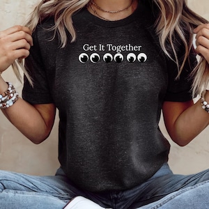 May include: A dark charcoal gray short-sleeve t-shirt featuring the white text Get It Together above a graphic of six white and black googly eyes arranged in a horizontal row across the center of the chest area of the garment.