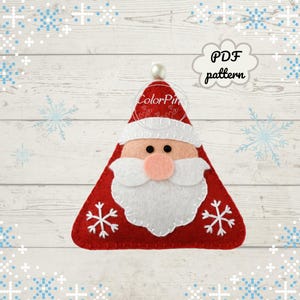 May include: A festive felt Christmas decoration in the shape of Santa Claus. The ornament is red with white detailing, a white beard, and a pink nose. Two white snowflakes adorn the bottom. The text "PDF pattern" is in a cloud-shaped bubble.