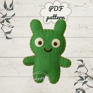 May include: A green felt toy creature with a smiling face, large eyes, and stitched details. The text "PDF pattern" is in a cloud-shaped speech bubble above the figure. The background is a light wood grain.