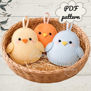 Può includere: Tre ornamenti a forma di pulcino in feltro in un cestino di vimini. I pulcini sono gialli, arancioni e azzurri, ognuno con occhi neri, becchi arancioni e nastri bianchi. L'immagine include il testo "PDF pattern".