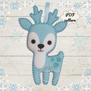 May include: A light blue felt deer ornament with white accents and black eyes. The deer has a ribbon for hanging and a decorative pattern on its body. The text "PDF pattern" is visible.
