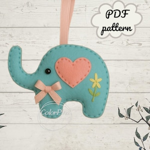 May include: A teal felt elephant ornament with a pink heart and bow. A yellow flower is stitched on the side. The ornament hangs from a pink ribbon. The words "PDF pattern" are in a cloud-shaped text bubble.