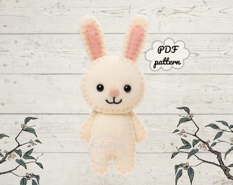 Può includere: Un coniglietto di feltro color crema con l'interno delle orecchie rosa, occhi neri e un sorriso cucito. Il coniglietto è in piedi. Il testo in una nuvola dice "PDF pattern". Lo sfondo è un legno bianco.