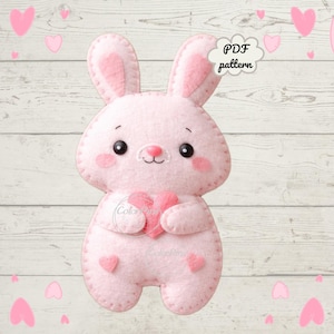 Puede incluir: Un conejito rosa de fieltro sosteniendo un corazón rosa, con ojos negros y una pequeña nariz rosa. El conejito tiene detalles rosas en las mejillas y las orejas. La imagen incluye el texto "PDF pattern" en una burbuja de diálogo en forma de nube.