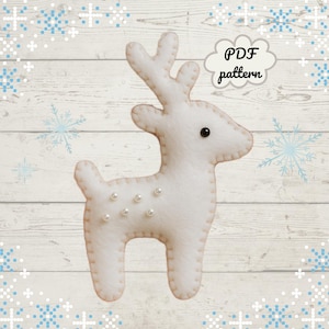 May include: A white felt reindeer with pearl accents and a black eye. The reindeer has a stitched outline and is standing on a light wood background with snowflake accents. Text in a cloud bubble reads "PDF pattern".