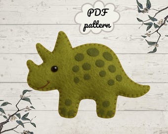 May include: A green felt dinosaur with dark green spots and a smiling face. The dinosaur has three horns and is standing on four legs. The text "PDF pattern" is in a cloud-shaped bubble above the dinosaur.