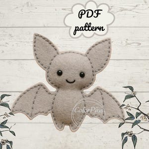 Peut inclure: Une chauve-souris en feutre gris clair avec des détails cousus, de grandes oreilles et des ailes déployées. La chauve-souris a un visage souriant avec des yeux noirs. Le texte "PDF pattern" est dans une bulle en forme de nuage. Fond en bois clair.