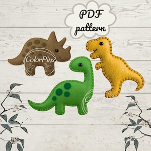 May include: Three felt dinosaur shapes: a brown triceratops with dark spots, a green brontosaurus with green spots, and a yellow T-Rex. The dinosaurs are stitched together with contrasting thread. The text "PDF pattern" is in a cloud-shaped speech bubble.