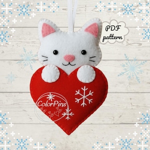 Puede incluir: Adorno de gato de fieltro blanco con orejas internas rosas y nariz rosa, sosteniendo un corazón rojo. El corazón tiene un copo de nieve y el texto "ColorPins" con un diseño de copo de nieve. El texto "PDF pattern" también es visible.