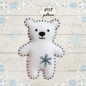 Può includere: Un orso polare di feltro bianco con dettagli cuciti neri e un abbellimento a forma di fiocco di neve. L'orso ha naso e occhi neri. L'immagine include il testo "PDF pattern" in una nuvoletta.
