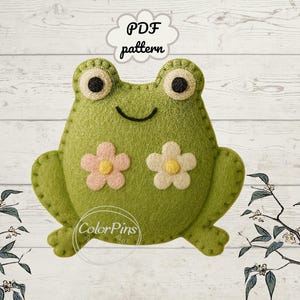 May include: A handmade green felt frog with large, round eyes and a smiling mouth. Two flower appliqués decorate the frog's chest. The image includes the text "PDF pattern" in a cloud-shaped bubble.