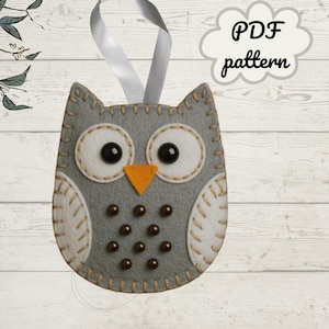 May include: A grey felt owl decoration with white detailing, black button eyes, and an orange beak. The owl has a stitched outline and a grey ribbon for hanging. The text "PDF pattern" is in a cloud-shaped speech bubble.