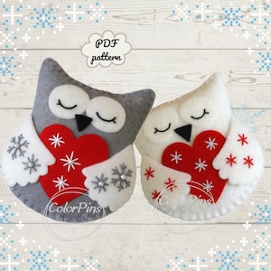 Peut inclure: Deux ornements en feutre en forme de hibou, un gris et un blanc, chacun tenant un cœur rouge. Les hiboux ont les yeux fermés et des accents de flocons de neige et d'étoiles. Le texte "PDF pattern" est dans un nuage au-dessus des hiboux.