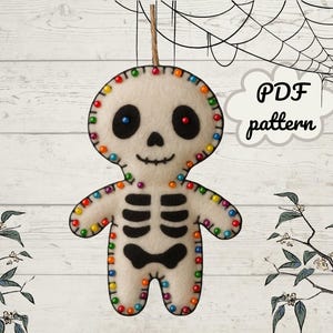 May include: A felt skeleton ornament with a smiling face, black eyes, and a black bow tie. The skeleton is decorated with colorful beads around the edges and along the rib cage. The text "PDF pattern" is in a cloud-shaped speech bubble.