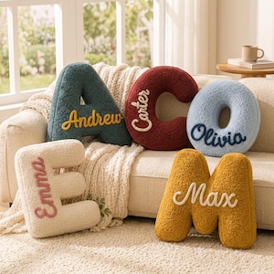 May include: Five plush letter-shaped pillows made of textured sherpa fabric. The dark teal letter A features the name Andrew, the maroon C features Carter, the light blue O features Olivia, the cream E features Emma, and the mustard M features Max.