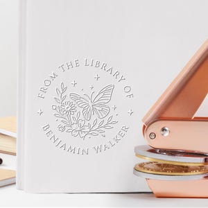 May include: A white book with a debossed floral and butterfly design, and the text "FROM THE LIBRARY OF BENJAMIN WALKER." A rose gold book embosser is in the foreground. Additional books and a pen are in the background.