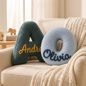 May include: A dark teal letter A pillow with yellow embroidered text reading Andre and a light blue letter O pillow with dark blue embroidered text reading Olivia. Both pillows are made of a textured, fuzzy, sherpa-like fabric material.