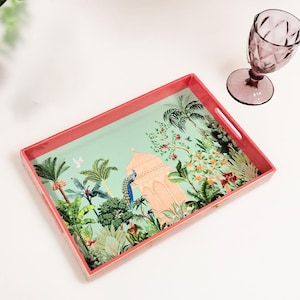 Puede incluir: Una bandeja rectangular con marco color coral y un interior verde claro que presenta una ilustración impresa de un edificio color melocotón, palmeras y un pavo real. Una copa de cristal morada con textura facetada se sitúa al lado.