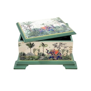 Può includere: Una scatola rettangolare in legno con base e coperchio verde salvia, caratterizzata da una scena dipinta di elefanti blu con selle e cavalieri colorati tra palme e fogliame. La scatola ha un coperchio incernierato e quattro piedini.