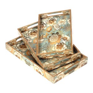Op de afbeelding: Een set van vier decoratieve houten serveerbladen. De bladen hebben een bloemenontwerp met gouden en witte bloemen op een lichtblauwe achtergrond. Elk blad heeft een rechthoekige vorm met handvatten.