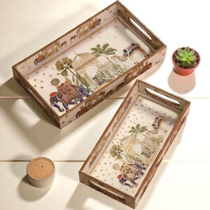 Puede incluir: Dos bandejas decorativas rectangulares con asas. Las bandejas presentan un fondo crema con lunares dorados y una ilustración detallada de elefantes, camellos y elementos arquitectónicos. Las bandejas tienen un marco de madera.