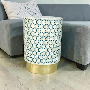 May include: A cylindrical side table featuring an intricate geometric pattern of white and teal inlaid tiles across its body. The base of the table is constructed from a solid, smooth gold-toned metal, providing a stable foundation for the piece.