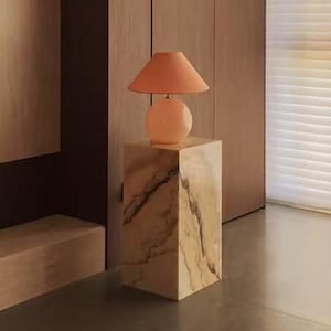 Puede incluir: Una lámpara de mesa con base redonda y pantalla cónica en tono melocotón suave, apoyada sobre un pedestal rectangular de mármol con vetas marrones marcadas. La lámpara se sitúa en una estancia con paneles de madera oscura y una silla capitoné de color rojo intenso.