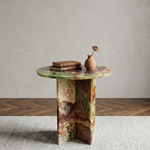May include: A round side table crafted from multicoloured onyx stone featuring a polished surface with swirling patterns of green, brown, and purple. The table has a cross-shaped base and supports two stacked brown books and a small terracotta vase.