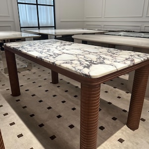 Op de afbeelding: Een rechthoekige tafel met een marmeren blad met wit en grijs geaderd patroon. Het frame en de poten zijn van bruin hout met groeven. Afmetingen: ongeveer 183 cm lang en 91 cm breed, ideaal voor eetkamer of tentoonstelling.