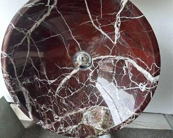 May include: A circular vessel sink made of polished deep red marble with prominent white veining throughout the stone surface. The basin features a centered, reflective chrome-finished drain assembly, showcasing a smooth, glossy finish and natural stone patterns.