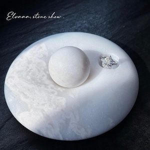 May include: A white marble ring holder with a pear-shaped diamond ring and a spherical accent. The holder has a circular design with a textured, marbled appearance. The text "Elvaaa. stone show." is in the upper left corner.